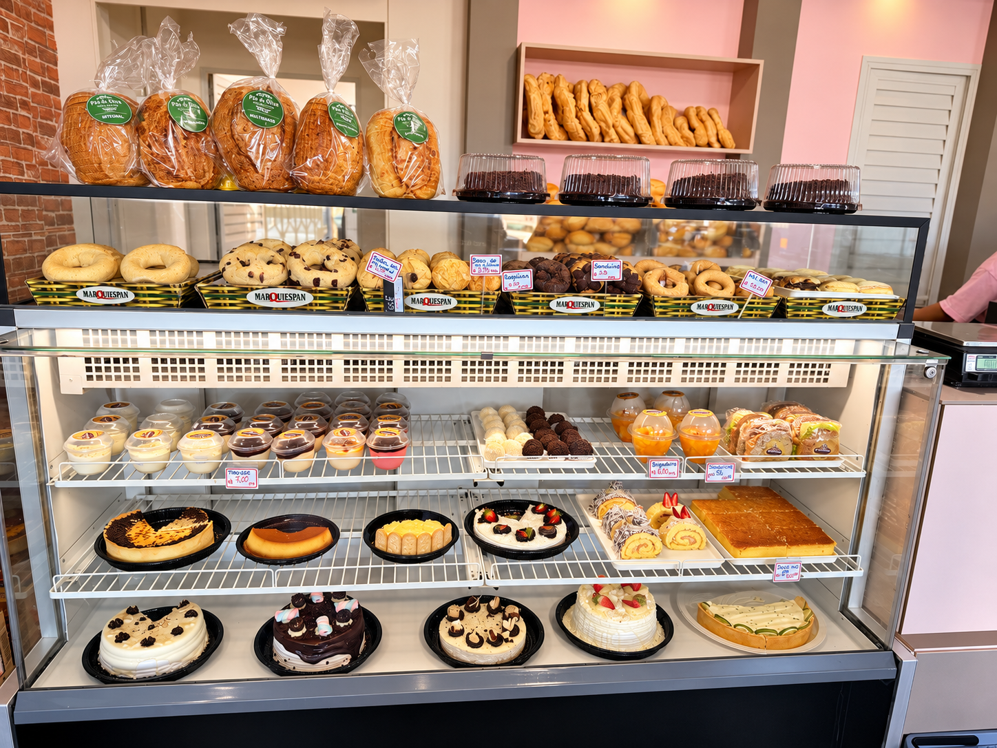 Balcão e vitrine da padaria com pães embalados, doces em cestas, sobremesas e bolos confeitados
